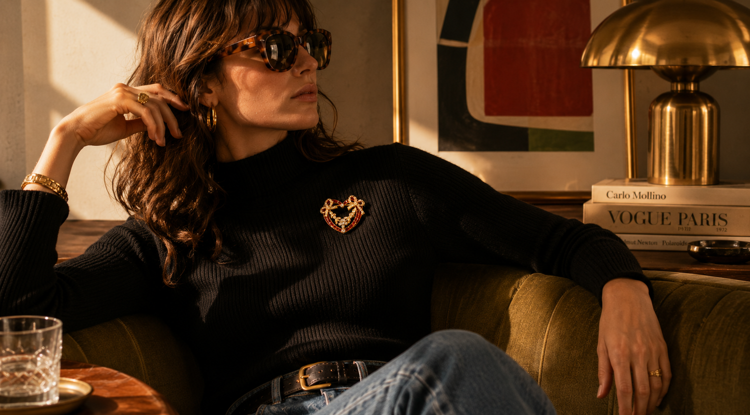 Woman sitting on a couch wearing sunglasses and a black sweater with a brooch, in a room with a lamp and books.